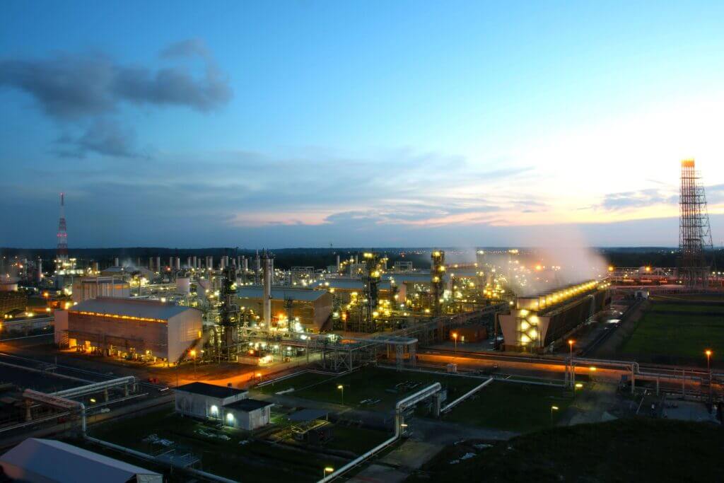 An industrial complex at dusk, with lights illuminating the sprawling network of pipes, towers, and buildings, and steam rising from cooling towers.
